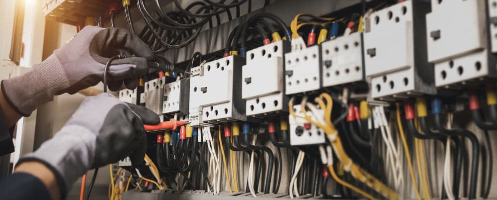 Electrician testing panel connections with multimeter, wearing gloves, in control panel.